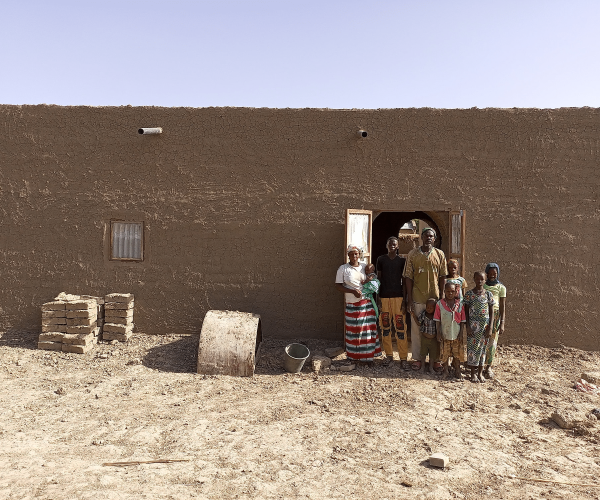 Une famille de neuf personnes, adultes et enfants, pose devant la porte d'une maison en terre neuve ; des briques sont empilées à gauche.