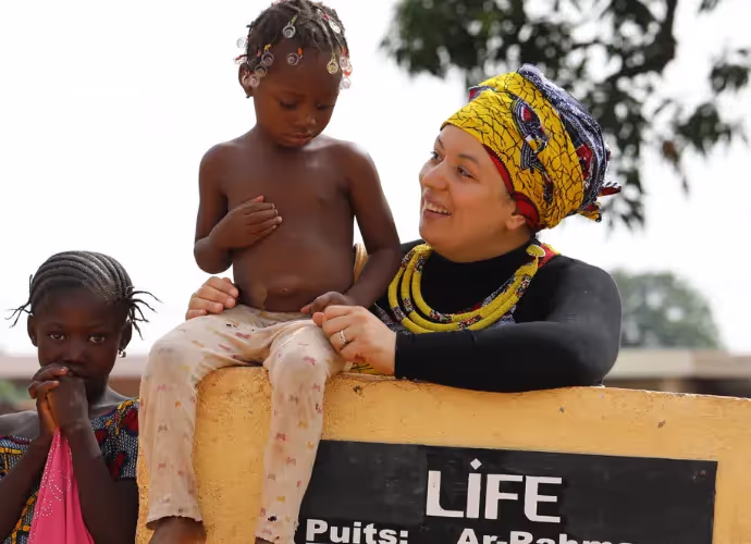 Une femme souriante en turban jaune tient un jeune enfant assis sur un muret portant l'inscription LIFE, Puits ; une fillette est à côté.