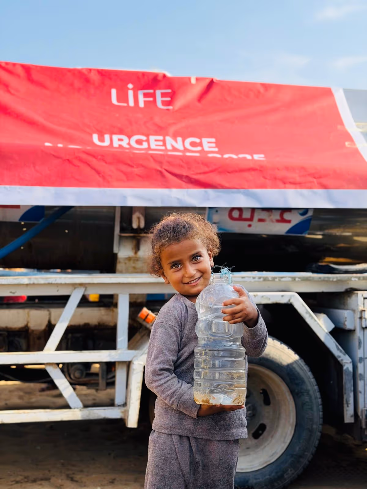 Petite fille souriante tenant une grande bouteille d'eau devant un camion-citerne avec une bâche rouge LIFE « Urgence »