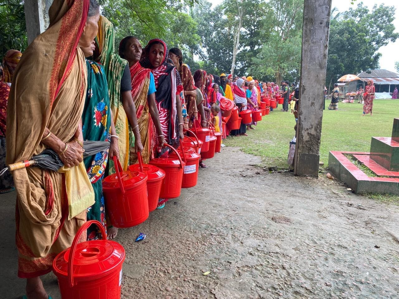 File de femmes en saris colorés, chacune avec un seau rouge à couvercle, attendant une distribution en plein air