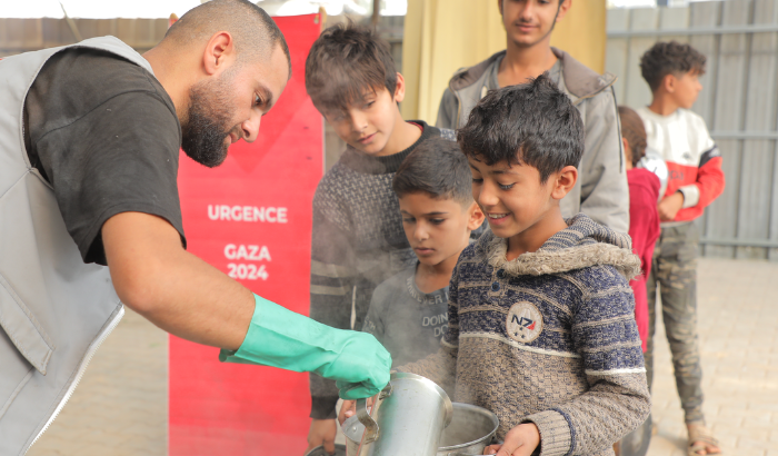 Un homme ganté sert à la louche un repas chaud à des enfants souriants, devant une bannière rouge « Urgence Gaza 2024 »