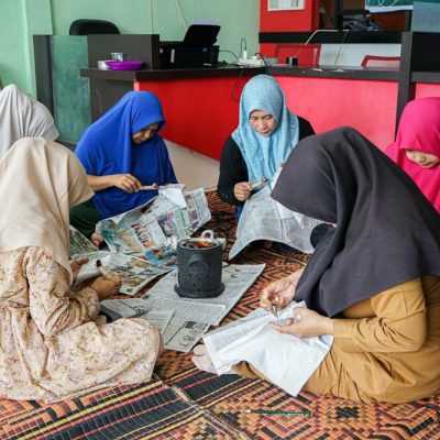 Des femmes voilées assises sur un tapis tressé travaillent du tissu blanc sur des feuilles de journal, autour d'un petit brasero