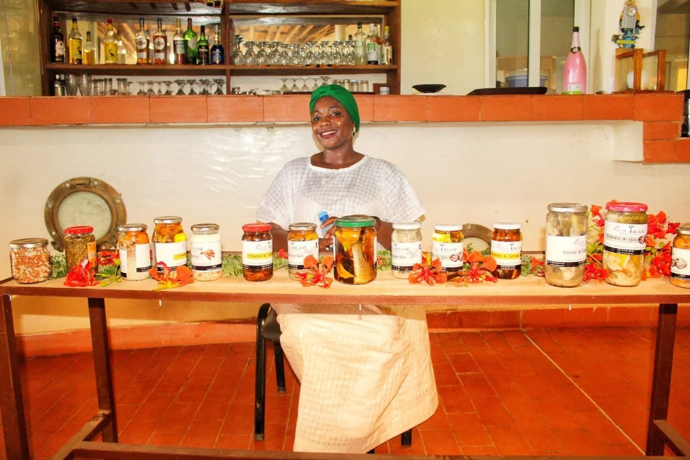 Femme souriante au foulard vert derrière une table alignant des bocaux de conserves artisanales ornés de fleurs rouges