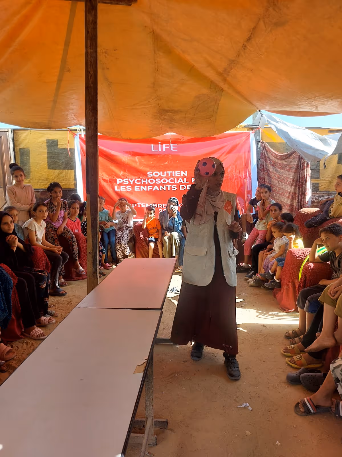 Une intervenante LIFE tient un ballon devant des enfants assis sous une tente, banderole « Soutien psychosocial pour les enfants »