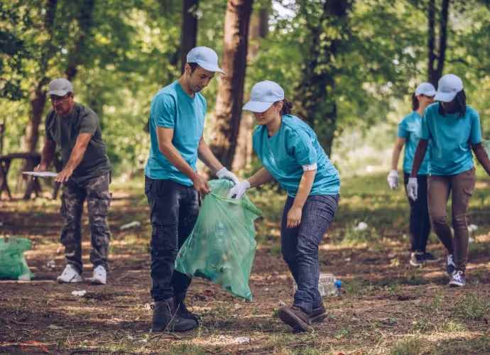Des bénévoles en t-shirts bleus et casquettes ramassent des déchets dans un sac vert en forêt