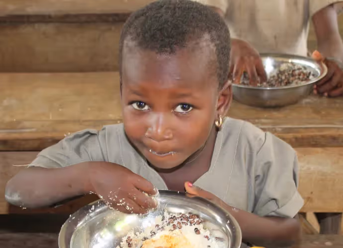 Jeune enfant mangeant du riz et des haricots avec la main dans une assiette en métal, devant un banc en bois