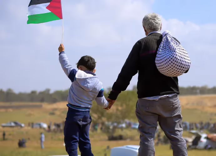 Un enfant tenant un drapeau palestinien lève le bras et donne la main à un homme âgé portant un baluchon, dans un champ.
