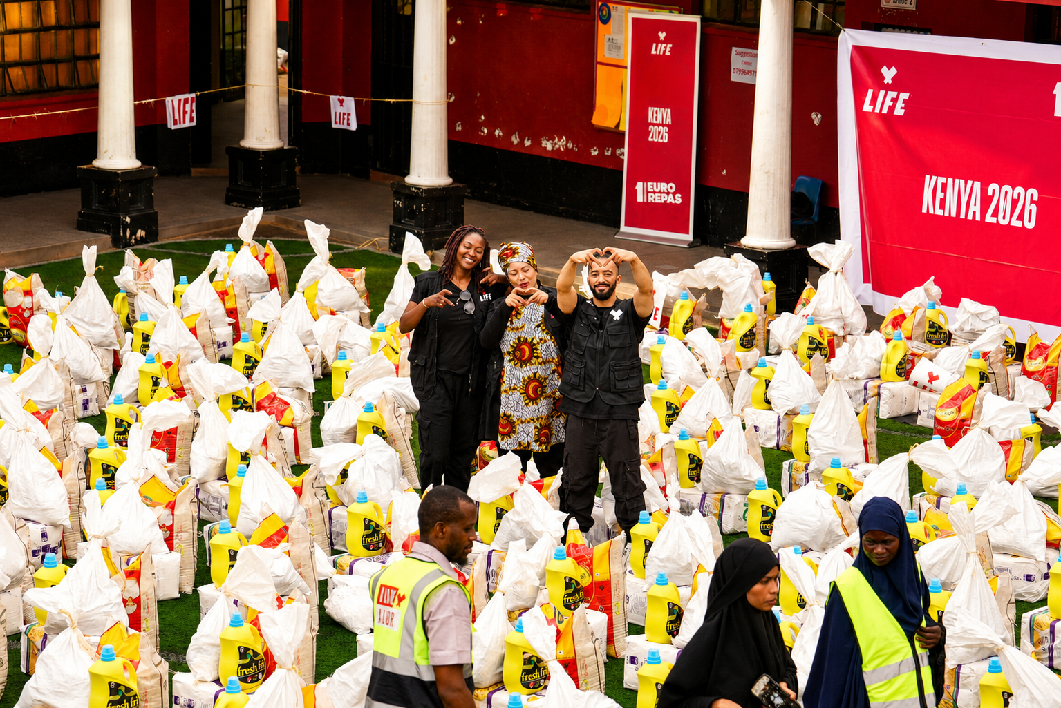 Trois personnes font un cœur avec les mains au milieu de sacs de riz et de bidons d'huile alignés, bannière LIFE Kenya 2026