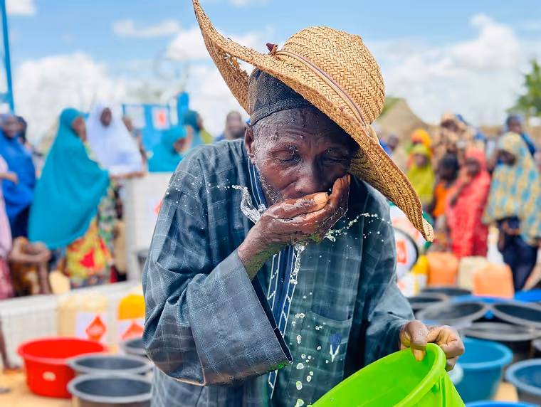 Un vieil homme coiffé d'un chapeau de paille boit de l'eau au creux de sa main, des bidons et une foule derrière lui.