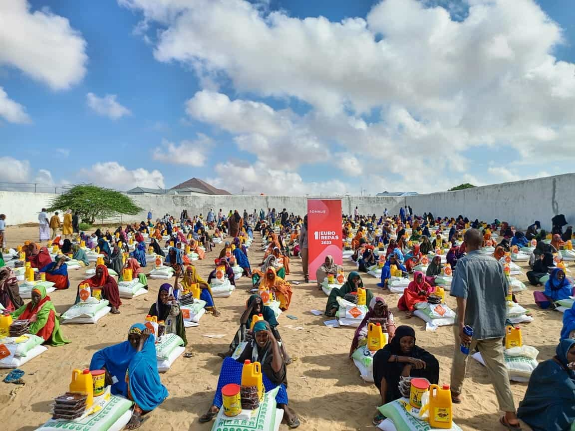 Distribution de colis alimentaires à de nombreuses femmes assises en rangs sur le sable, avec sacs et bidons d'huile, bannière « Somalie »