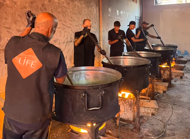Cinq hommes remuent de grandes marmites posées sur des feux dans un local ; l'un porte un gilet LIFE