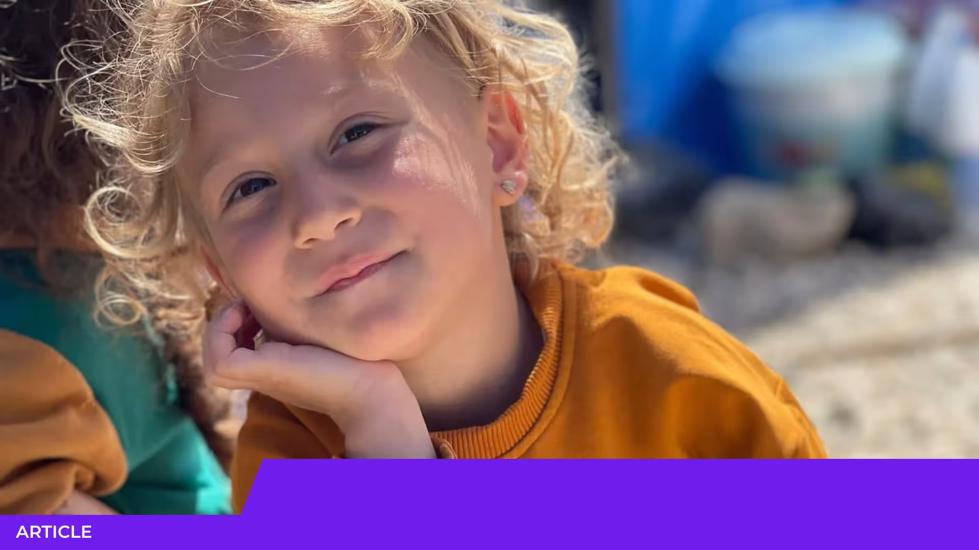 Portrait d'une jeune enfant blonde aux cheveux bouclés, en pull moutarde, la joue appuyée sur la main et souriante