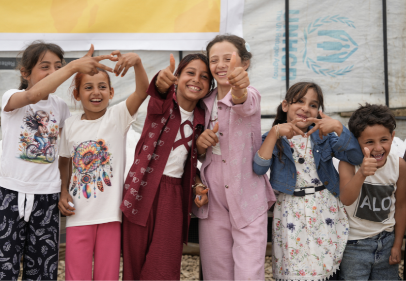 Groupe d'enfants souriants qui lèvent le pouce devant une bâche blanche portant le logo du HCR