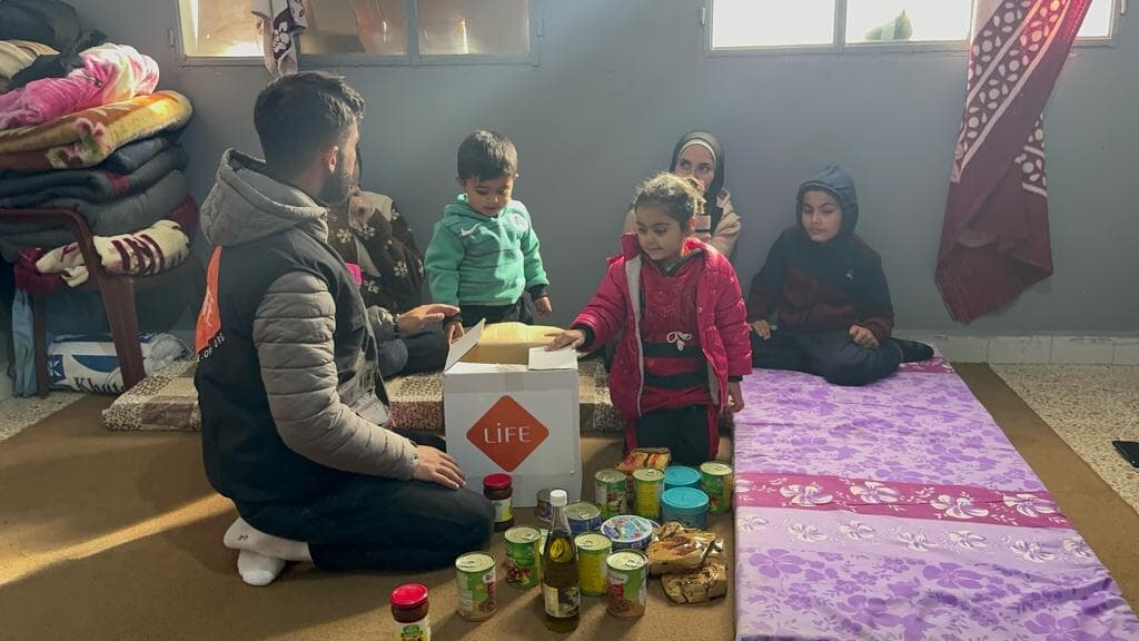 Un bénévole LIFE assis par terre distribue un colis de conserves et de provisions à des enfants dans une pièce