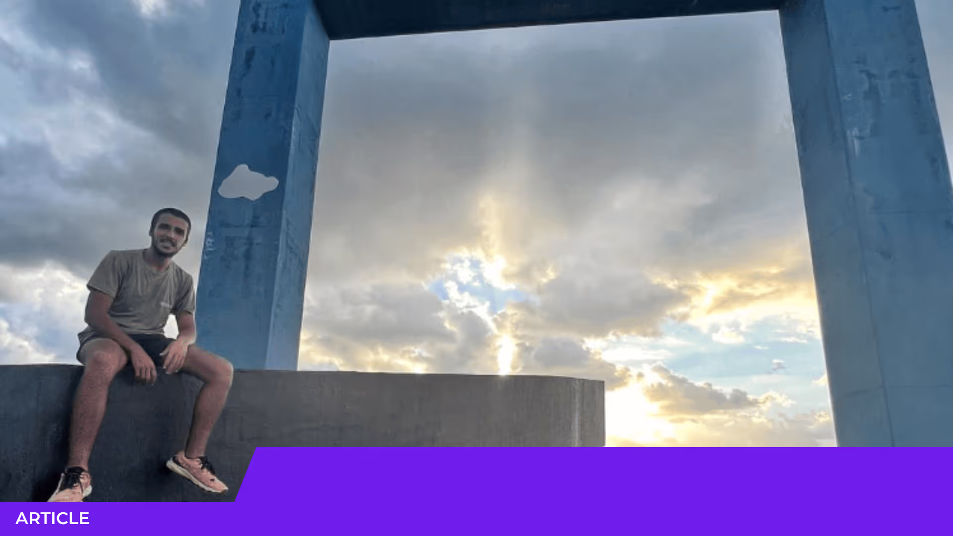Jeune homme souriant assis sur un muret en béton devant un grand cadre bleu, avec un ciel nuageux et des rayons de soleil