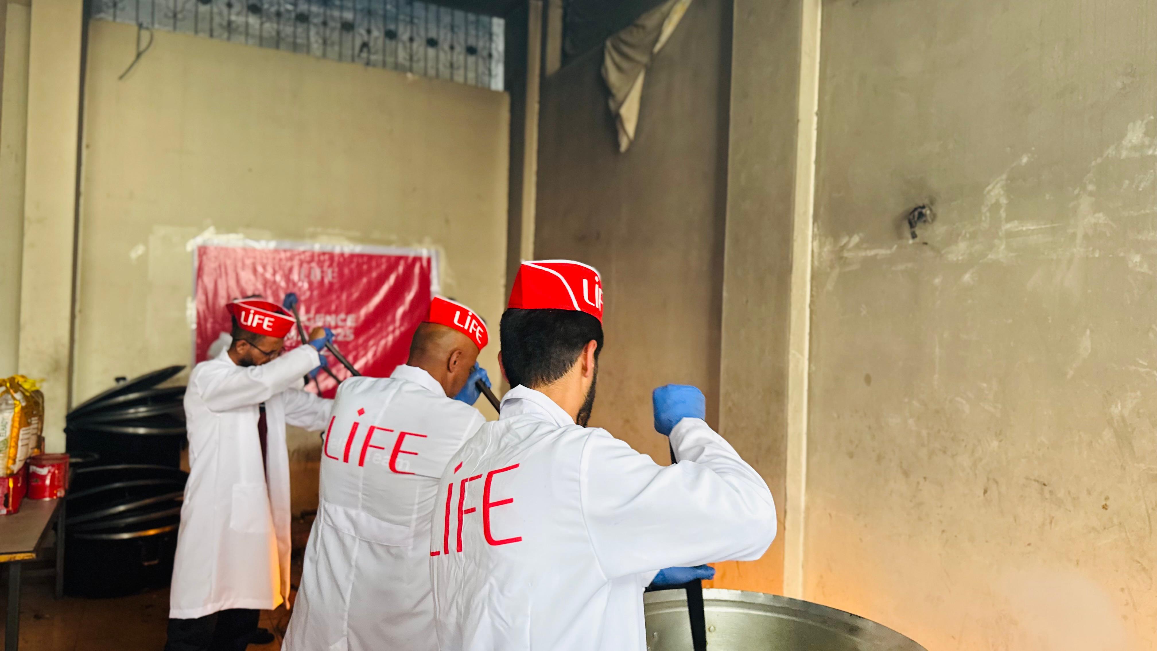 Trois bénévoles en blouse blanche et calot rouge LIFE remuent de grandes marmites dans une salle pour préparer des repas