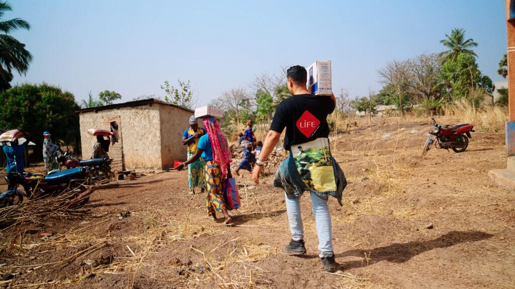 Un membre de LIFE en t-shirt noir porte un carton dans un village ; des femmes portent aussi des colis sur la tête, motos et case au fond.