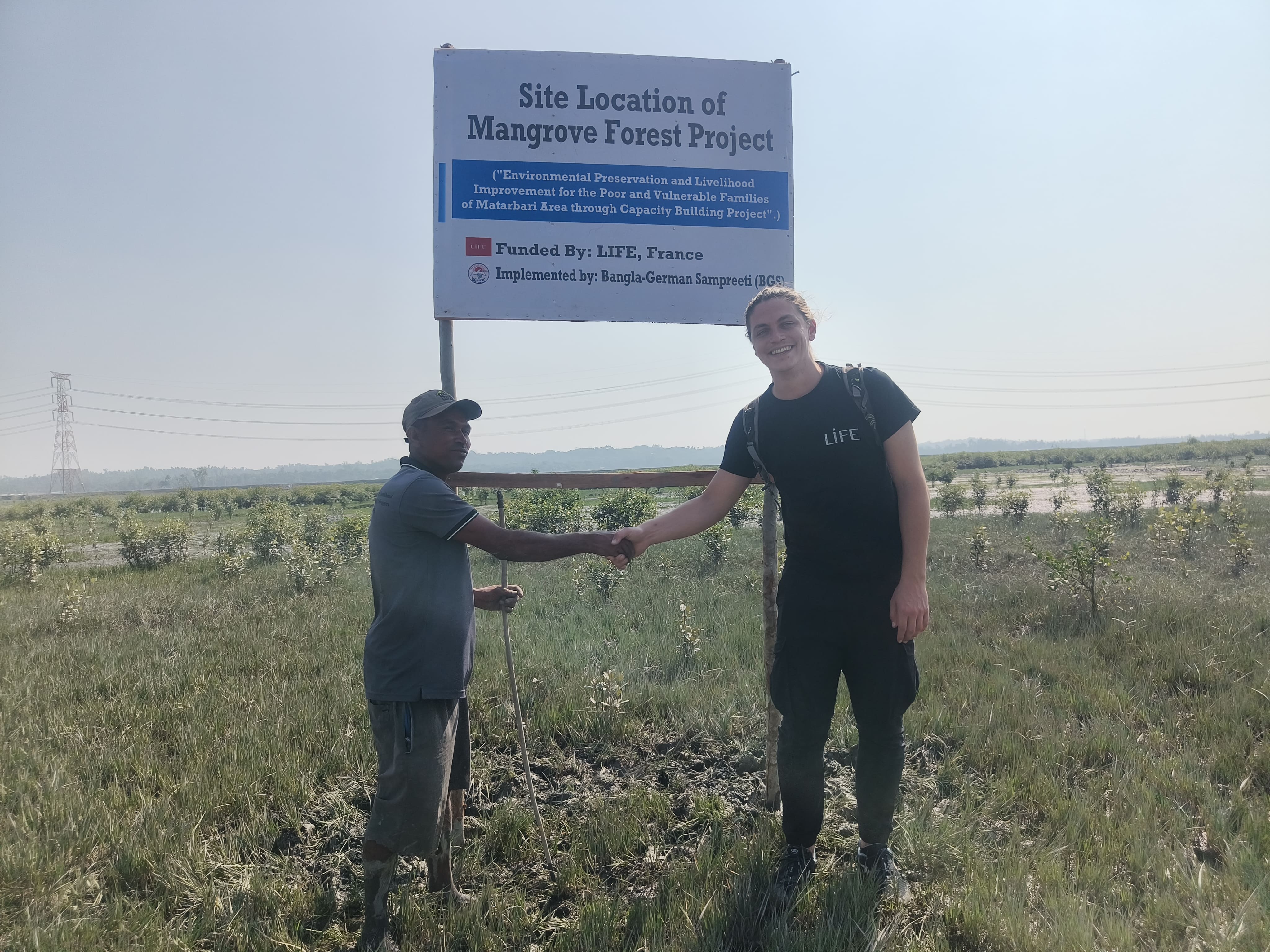 Deux hommes se serrent la main devant un panneau « Mangrove Forest Project » financé par LIFE, France, dans une plaine herbeuse