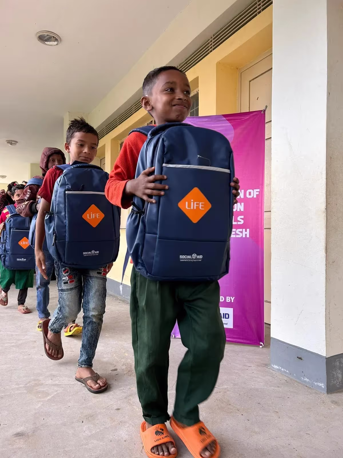 Écoliers en file dans une galerie d'école, portant sur le ventre des sacs à dos bleus ornés du logo LIFE