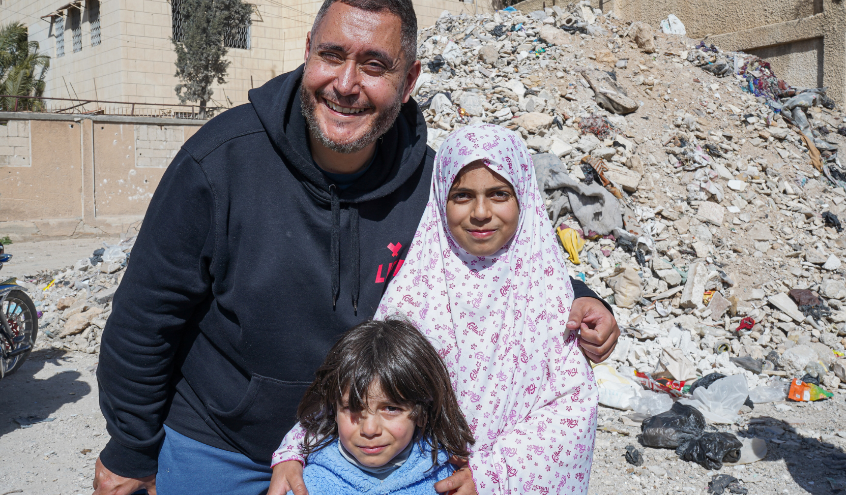 Homme souriant portant un sweat LIFE, entouré de deux enfants, devant un tas de gravats dans une rue
