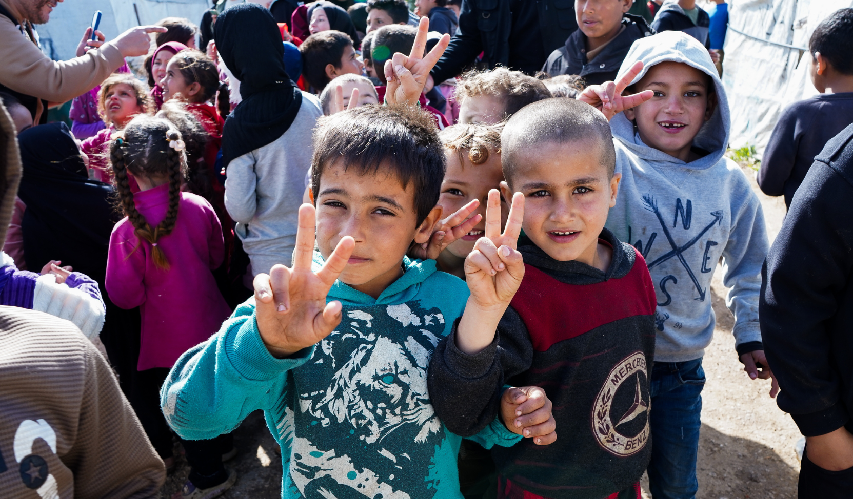Groupe d'enfants souriants dans un camp, deux garçons au premier plan faisant le signe de la victoire avec les doigts