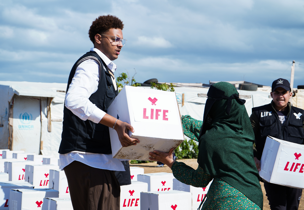 Un homme en gilet noir tend un carton LIFE à une femme en vert, devant des tentes et des piles de cartons.