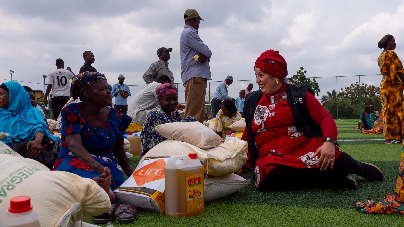Une responsable de LIFE assise sur l'herbe d'un terrain discute avec des femmes lors d'une distribution de sacs de riz et d'huile