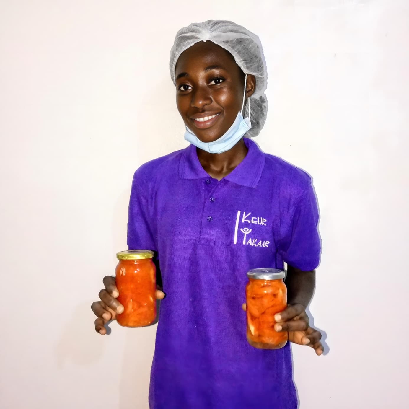 Jeune femme en polo violet Keur Takaar, charlotte sur la tête, souriante, tenant deux bocaux de préparation orangée.
