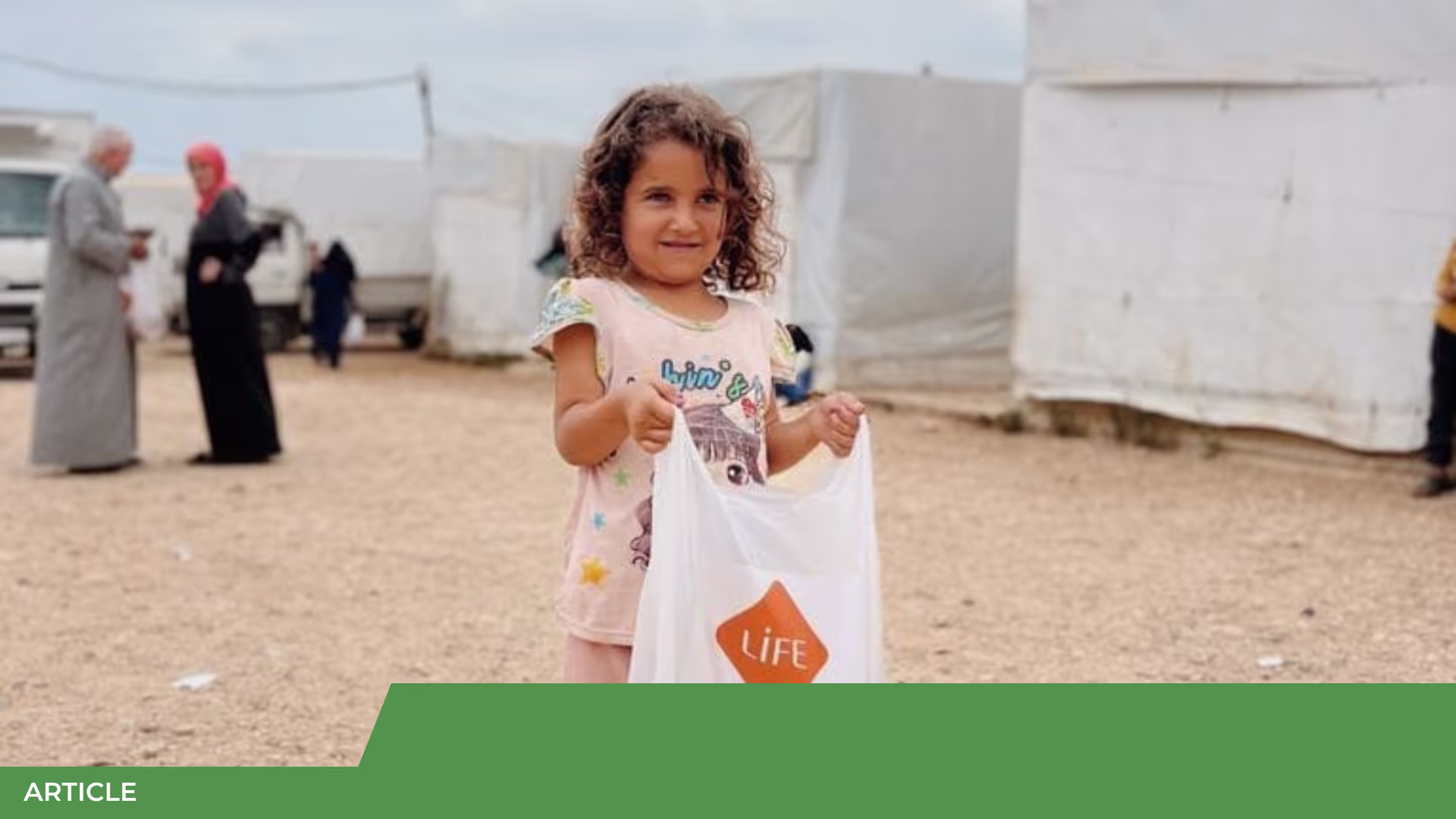 Fillette aux cheveux bouclés souriante tenant un sac blanc LIFE dans un camp de tentes, avec des adultes en arrière-plan