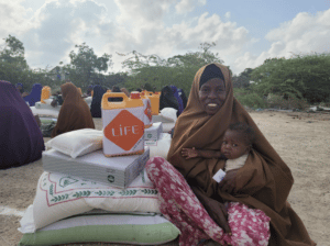 Femme voilée assise avec son bébé près de sacs de vivres et d'un bidon orange LIFE, lors d'une distribution alimentaire