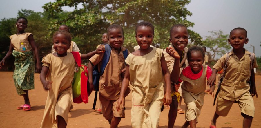 Groupe d'écoliers souriants en uniforme beige courant sur une piste de terre rouge, avec leurs sacs à dos