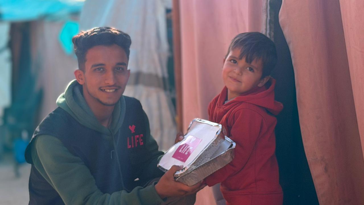 Un jeune homme en gilet LIFE tend une barquette repas à un petit garçon souriant en sweat rouge, devant des tentes.