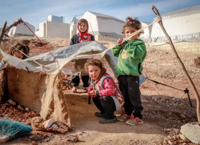 Trois jeunes enfants jouent près d'un abri de fortune en toile et en terre dans un camp de tentes, sur un sol caillouteux