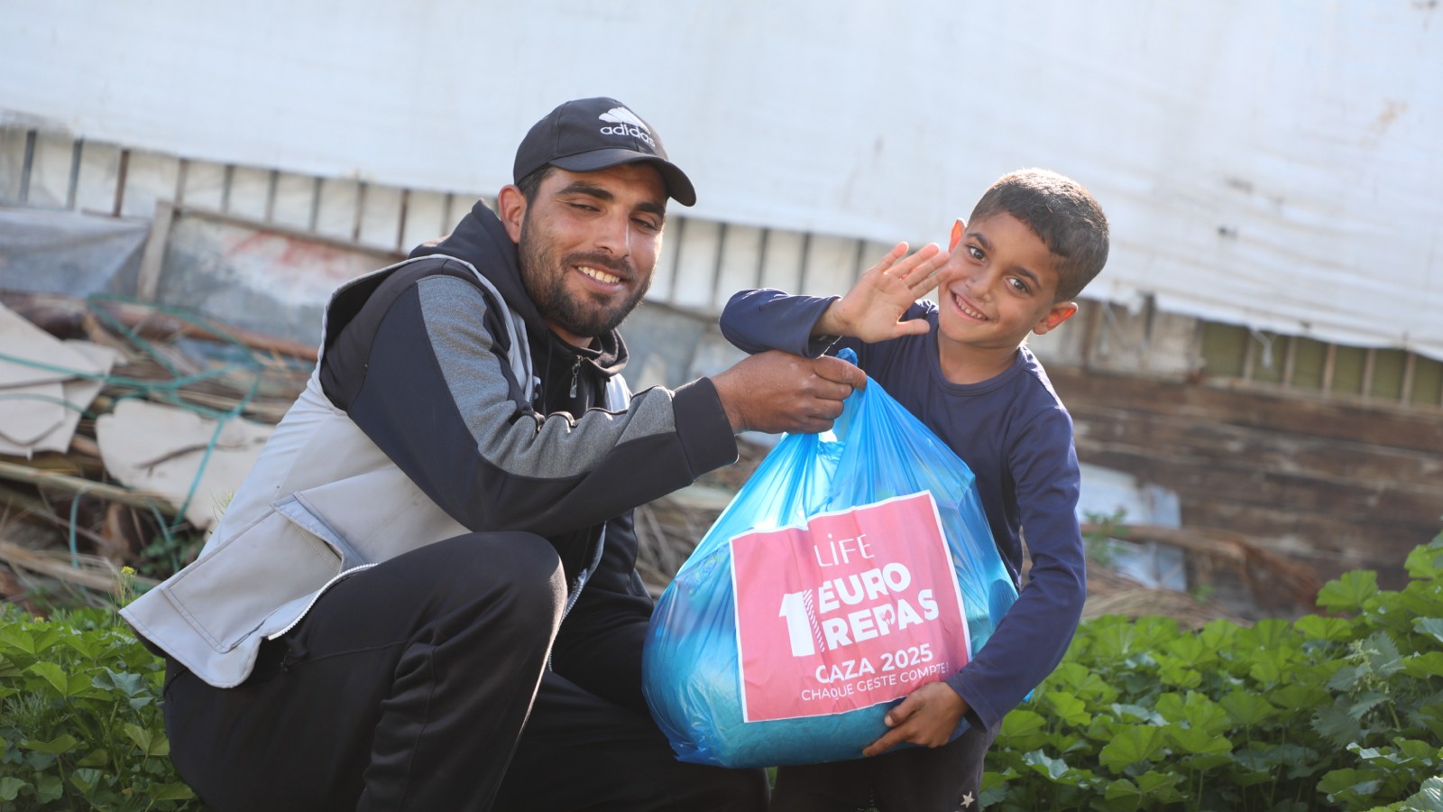 Un homme et un enfant souriants posent avec un sac bleu LIFE « 1 euro = 1 repas », Gaza 2025, l'enfant salue de la main
