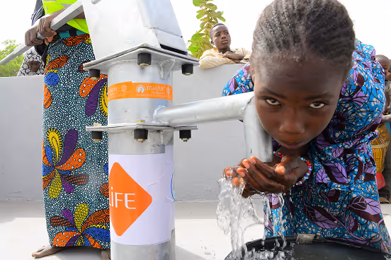 Une jeune fille boit l'eau d'une pompe manuelle portant le logo LIFE, dans ses mains jointes, devant d'autres personnes