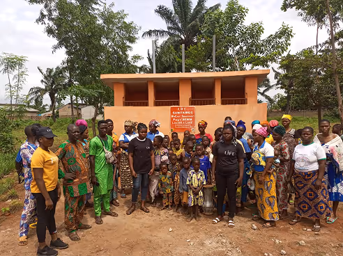 Groupe d'habitants et d'enfants, dont deux personnes en t-shirt LIFE, devant un bloc de sanitaires orange en construction