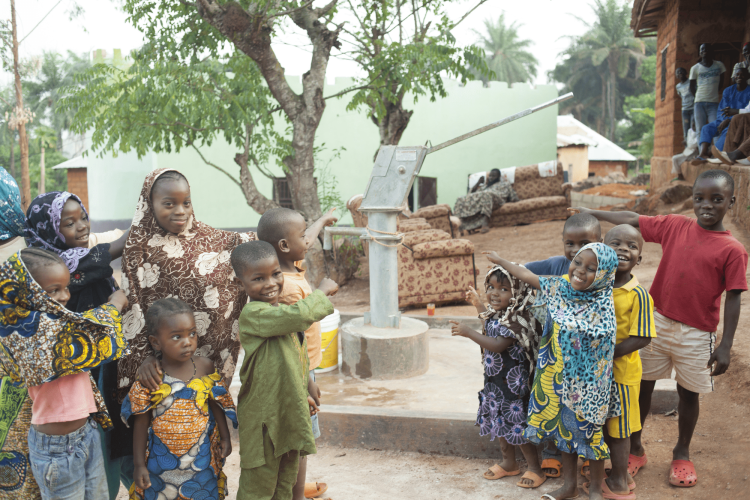 Groupe d'enfants souriants et de femmes rassemblés autour d'une pompe à main dans un village, des habitants assis au fond