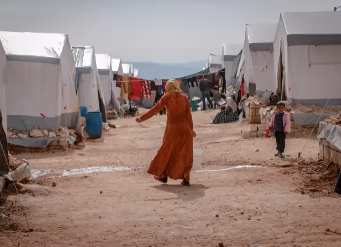 Une femme en robe orange marche dans une allée d'un camp de tentes blanches, un enfant se tient à droite