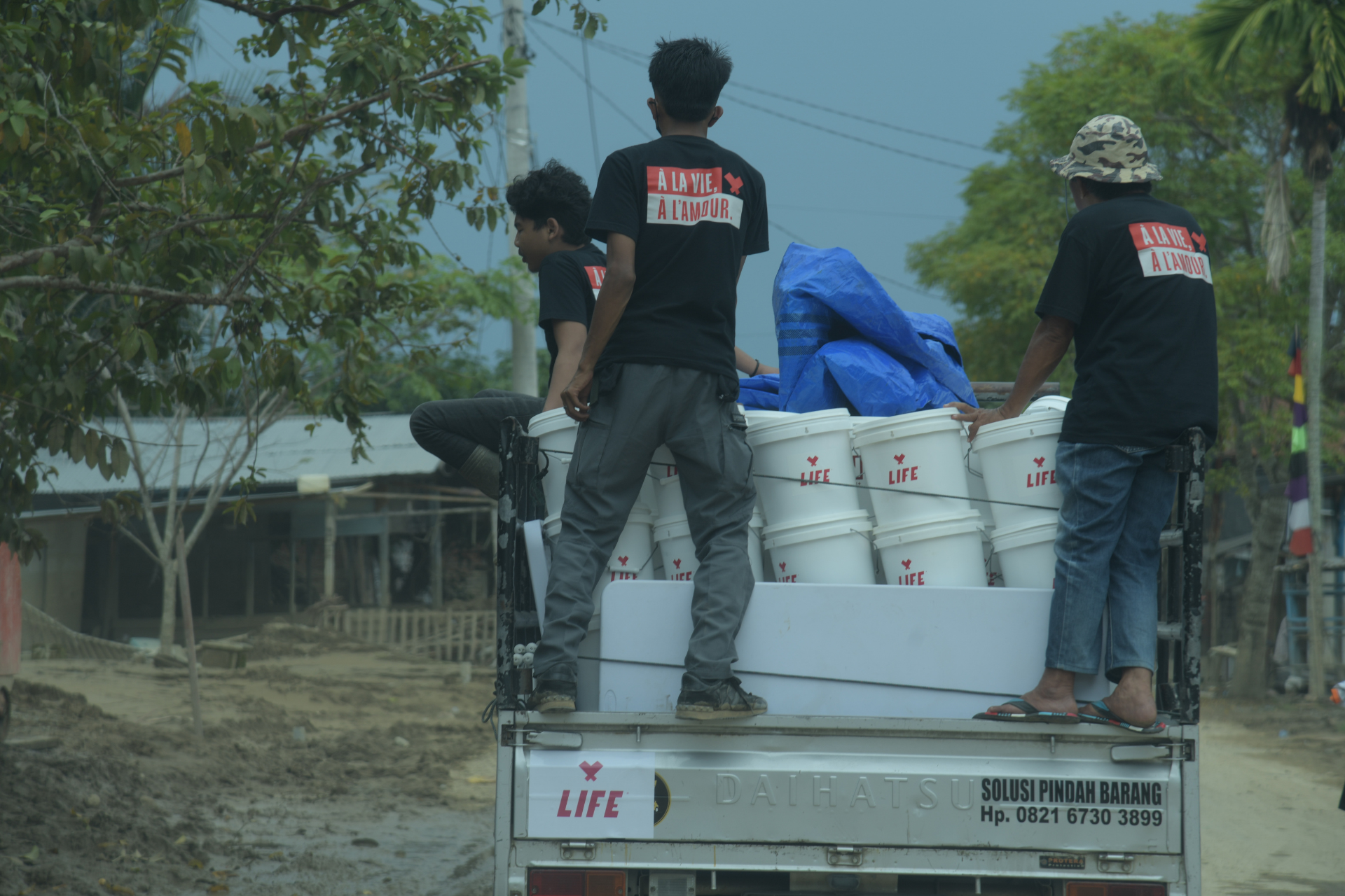 Des bénévoles en t-shirt « À la vie, à l'amour » chargent des seaux blancs LIFE à l'arrière d'un camion sur une route boueuse.