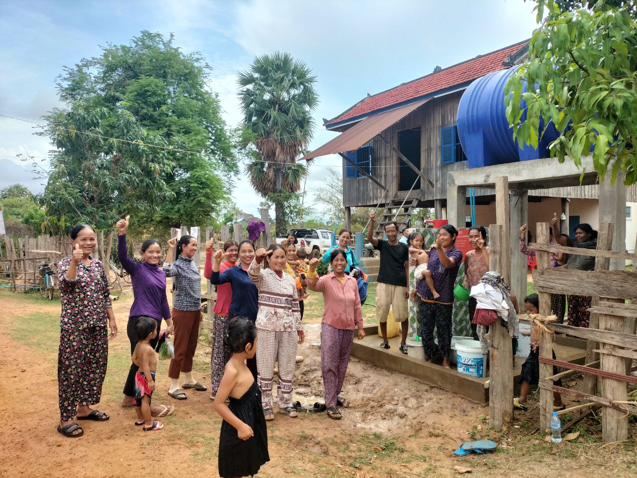 Des villageoises souriantes lèvent le pouce devant une maison sur pilotis équipée d'une grande citerne d'eau bleue