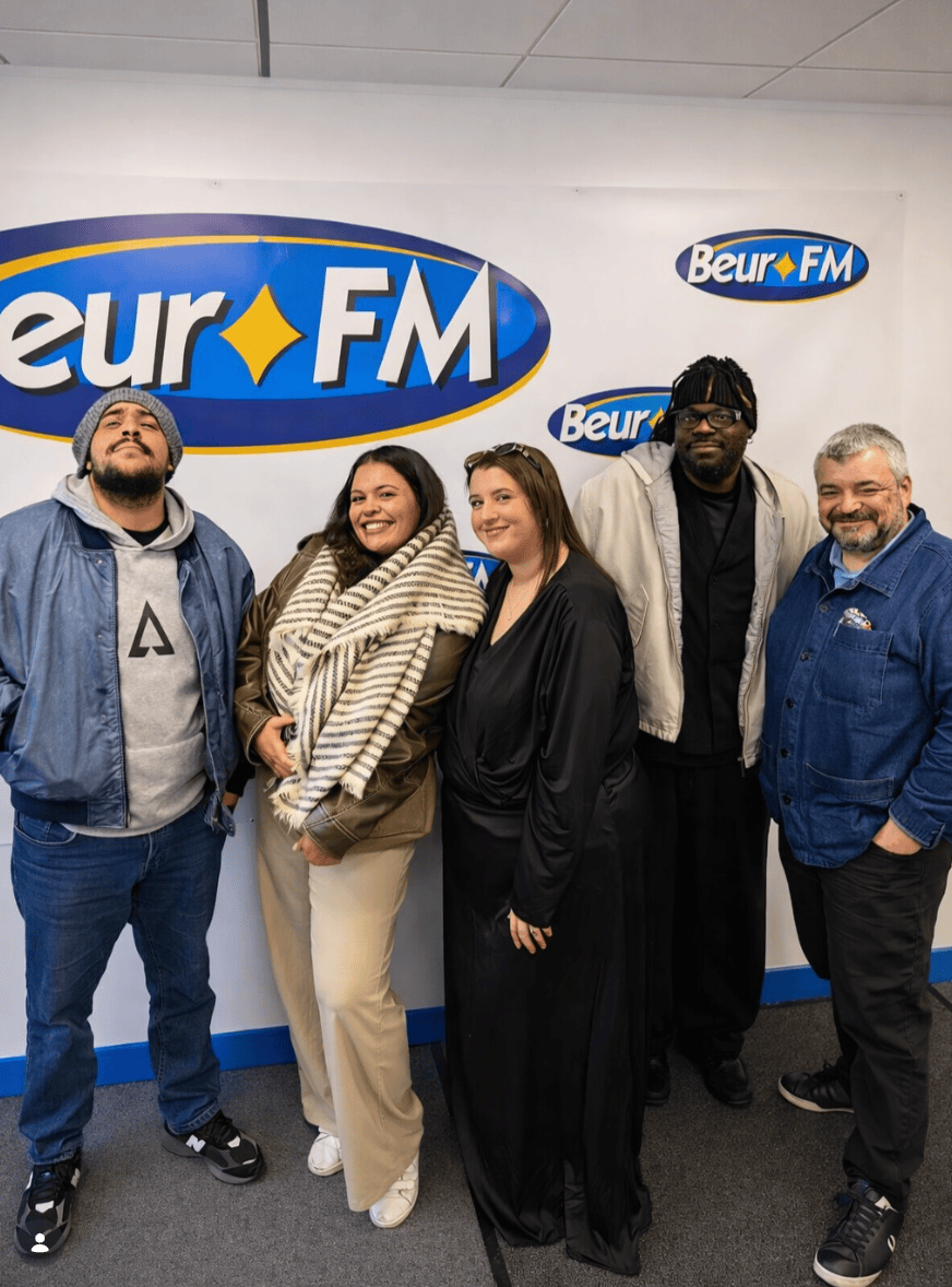 Cinq personnes posent côte à côte en souriant devant un mur blanc orné des logos de la radio Beur FM