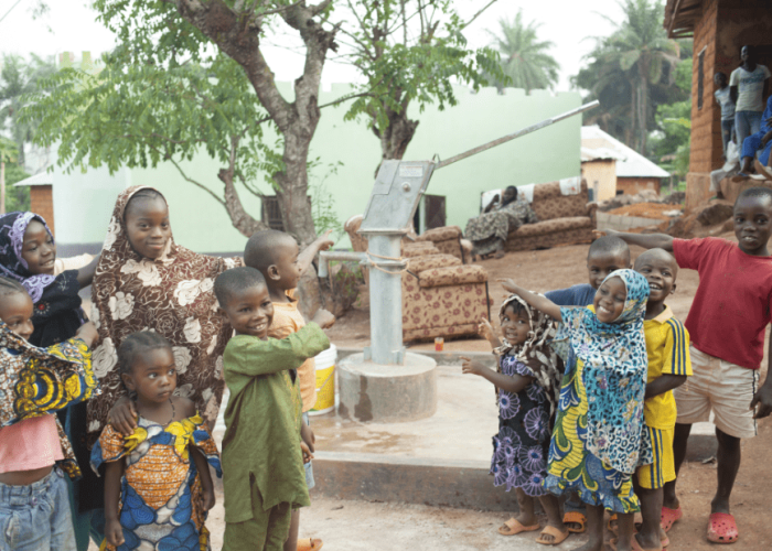 Groupe d'enfants souriants en tenues colorées devant une pompe à eau manuelle en béton, certains pointant du doigt la pompe