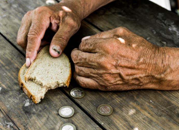 Mains burinées posées sur une table en bois, près de morceaux de pain et de quelques pièces de monnaie