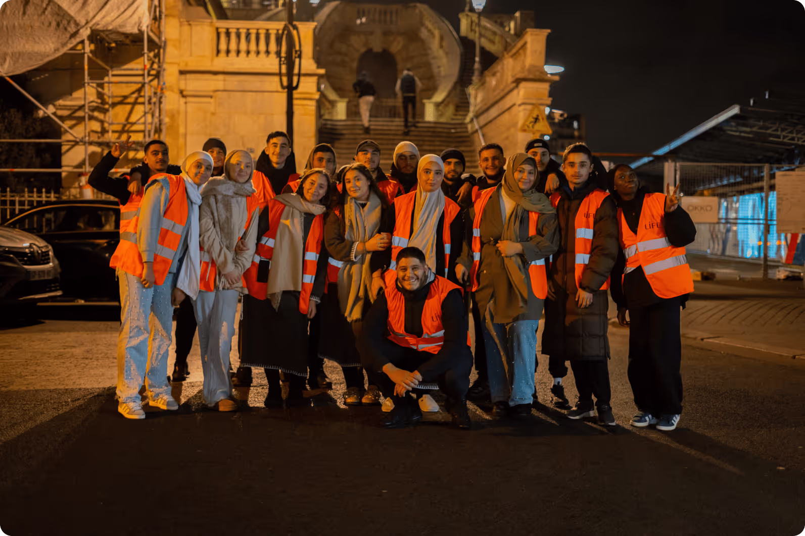 Groupe de jeunes bénévoles en gilets orange réfléchissants posant la nuit dans une rue, devant un escalier en pierre