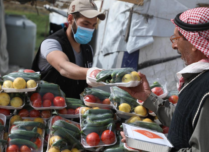 Un bénévole portant un masque et un gilet LIFE tend une barquette de légumes frais à un homme âgé coiffé d'un keffieh