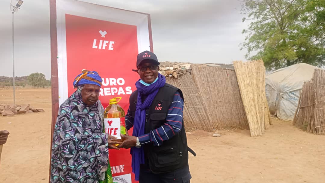 Un homme de LIFE en casquette remet une bouteille d'huile LIFE à une femme âgée, devant une bannière LIFE et des abris de paille