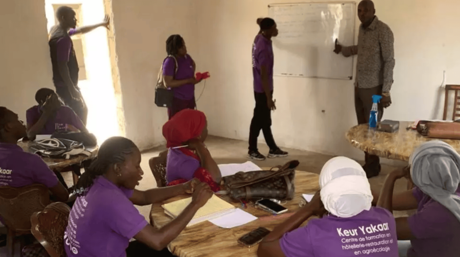 Salle de cours où des femmes en t-shirt violet Keur Yakaar suivent une formation, un formateur écrit au tableau blanc