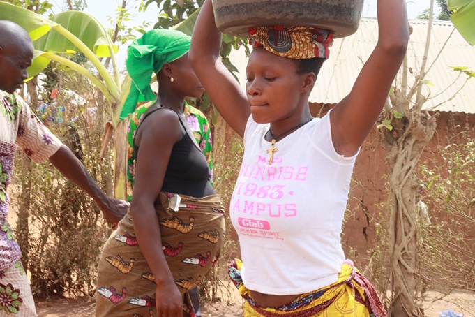 Une jeune femme porte une bassine sur la tête, les bras levés, près d'autres femmes dans un village