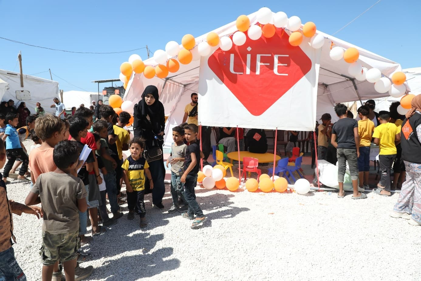 Des enfants font la queue sous une tente ornée de ballons orange et blancs, portant le logo LIFE, dans un camp