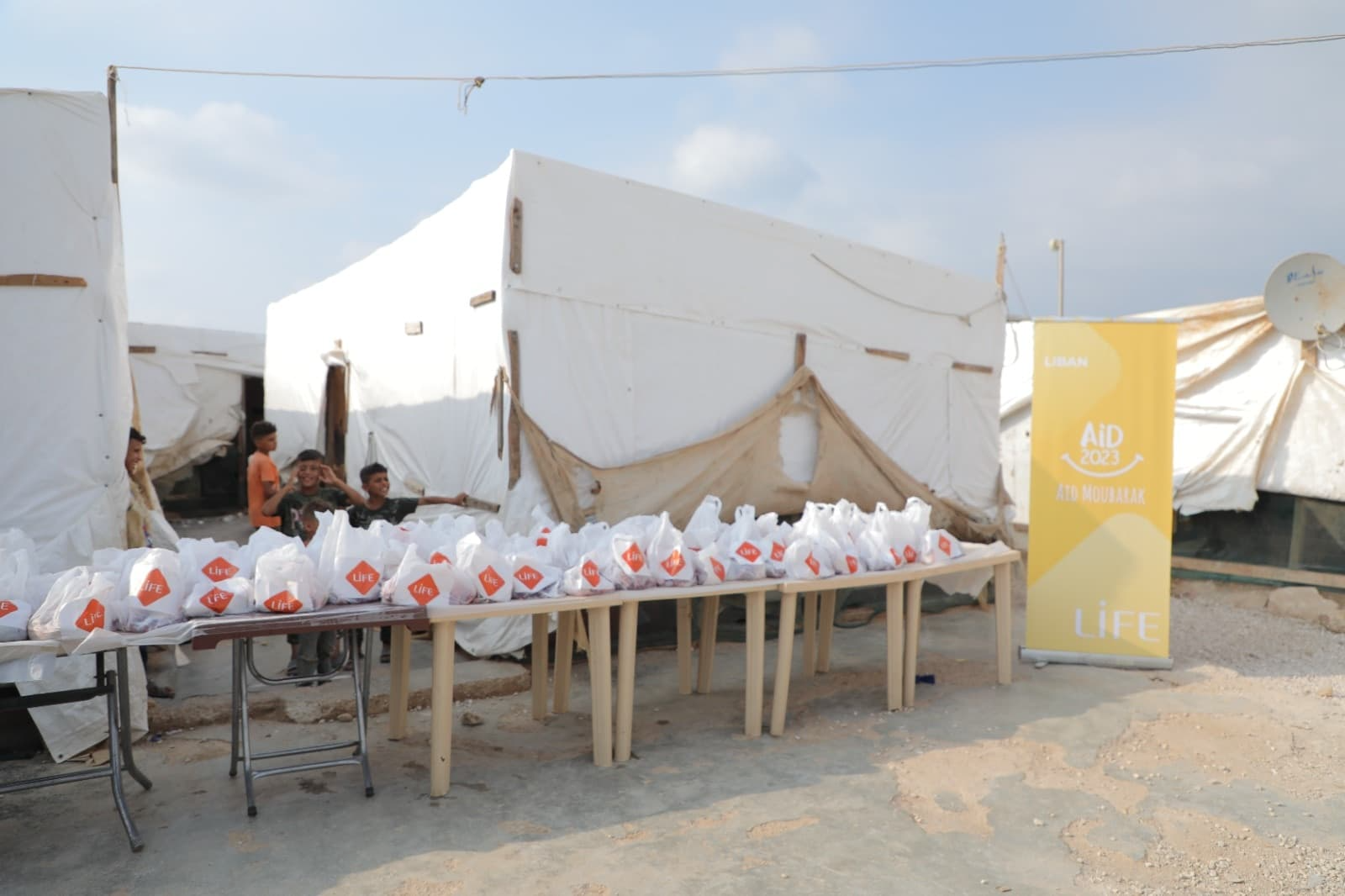 Table dressée devant des tentes avec des sacs blancs siglés LIFE alignés pour une distribution, bannière « Aïd Moubarak » au Liban